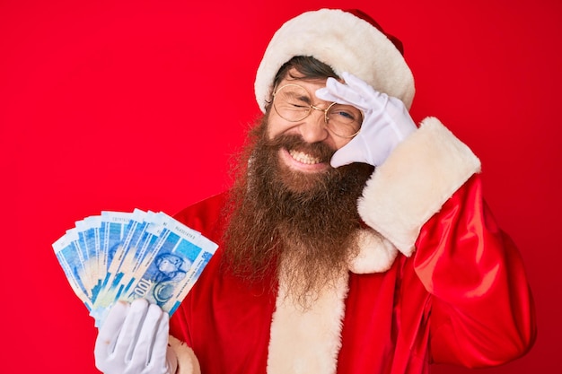 Bonito jovem ruivo com barba longa vestindo fantasia de Papai Noel segurando rand africano estressado e frustrado com a mão na cabeça, rosto surpreso e zangado