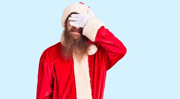 Bonito jovem ruivo com barba longa vestindo fantasia de Papai Noel sorrindo e rindo com a mão no rosto cobrindo os olhos de surpresa. conceito cego.