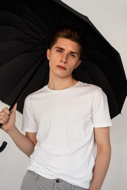 Bonito joven bastante serio en camiseta juvenil casual de moda se encuentra con paraguas vintage negro sobre fondo blanco en estudio. Chico guapo en ropa de moda con paraguas está aislado en la habitación.