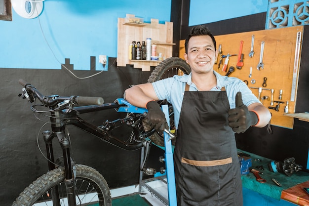 Bonito mecânico de bicicletas com avental, sorrindo, usando luvas e segurando a chave de boca com o polegar para cima