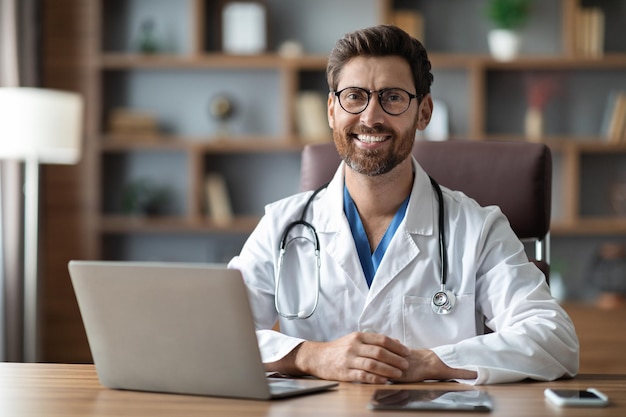 Bonito médico masculino de uniforme sentado na mesa com laptop no local de trabalho