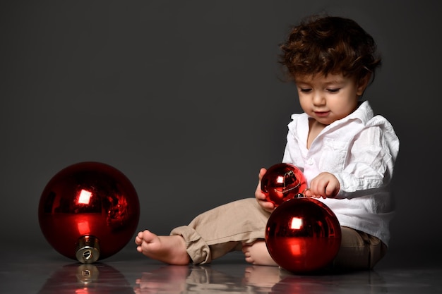 Bonito menino gordinho descalço bonito com roupa da moda brincando com bolas de bugiganga de brinquedo de Natal de tamanho diferente Retrato isolado filmado no fundo cinza do estúdio Feriado de inverno Conceito de véspera de Natal