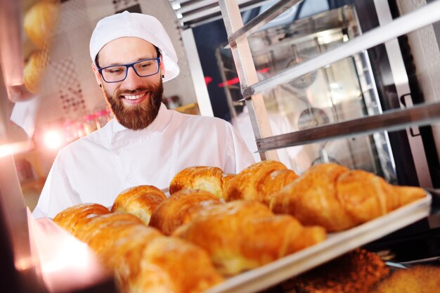 Bonito, padeiro, em, uniforme branco, segurando, em, seu, mãos, um, bandeja, cheio, de, freshly, assado, croissants