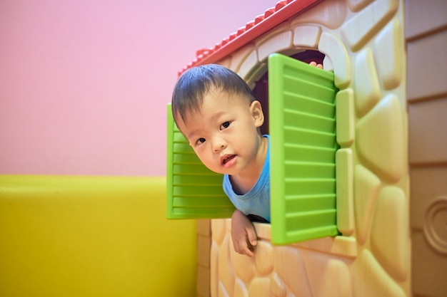 Bonito pequeno asiático 2 anos de idade criança menino está brincando de esconde-esconde na janela da casa de brinquedo de plástico,