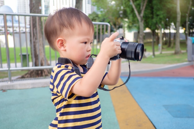 Bonito pequeno asiático 2 anos de idade menino da criança usar alça da câmera tirando foto usando a câmera digital, olhando para a câmera no parque, garoto fotografando a natureza, explorar e apreciar a natureza com o conceito de crianças