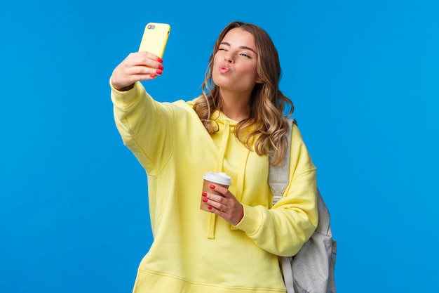 Foto bonito positivo e feminino bela jovem estudante feminino faculdade segurando o copo de papel de café favorito e mochila com livros, tendo selfie show beijo mwah rosto no celular