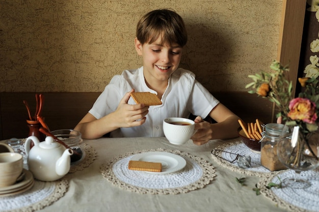 Bonito rapaz adolescente feliz sentado em casa à mesa e bebendo chá com biscoitos