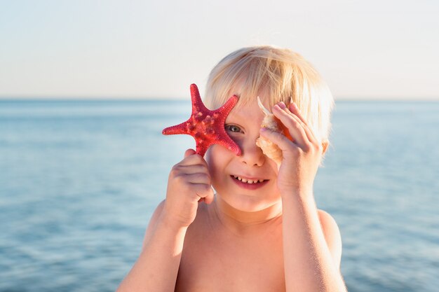 Bonito rapaz loiro detém estrela do mar e conchas e sorrindo.