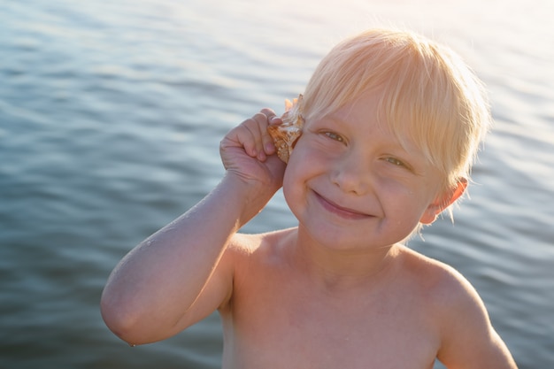 Bonito rapaz loiro, segurando a concha no ouvido. Parede do mar.