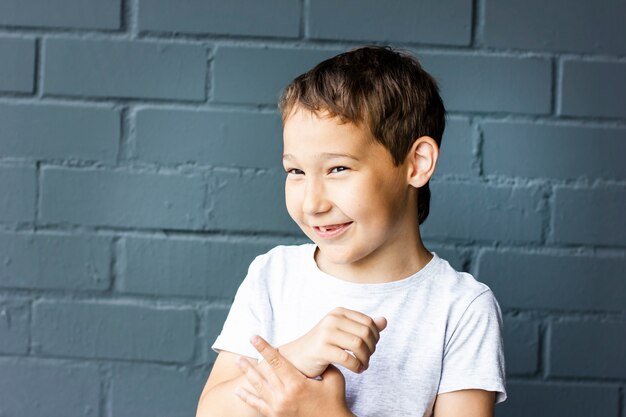 Bonito rapaz sorridente de 8 anos de idade confuso sobre o fundo da parede de tijolo cinza