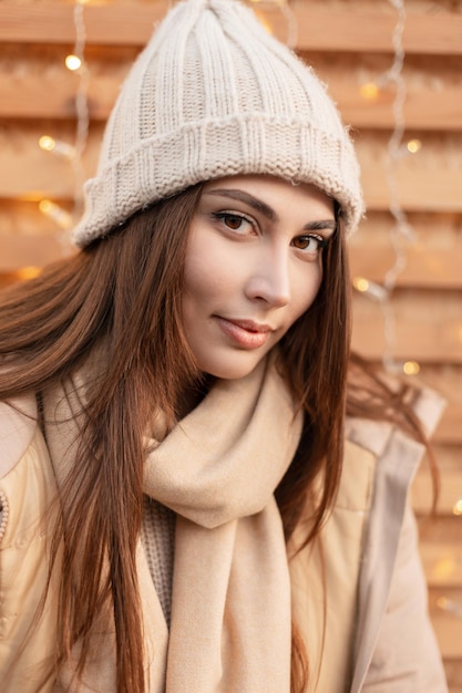 Bonito retrato de um modelo muito jovem em roupas da moda com um chapéu de malha vintage, jaqueta de inverno e cachecol Olha para a câmera perto de uma parede de madeira com luzes na rua