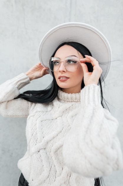 Bonito retrato femenino de hermosa joven con sombrero de mujer con suéter de punto de moda que usa y se pone unas gafas elegantes en la calle