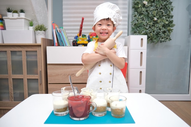 Bonito sorridente asiático menino de 4 anos de idade com um rolo se divertindo cozinhar bolo de morango em casa, diversão atividades internas para o conceito de jardim de infância