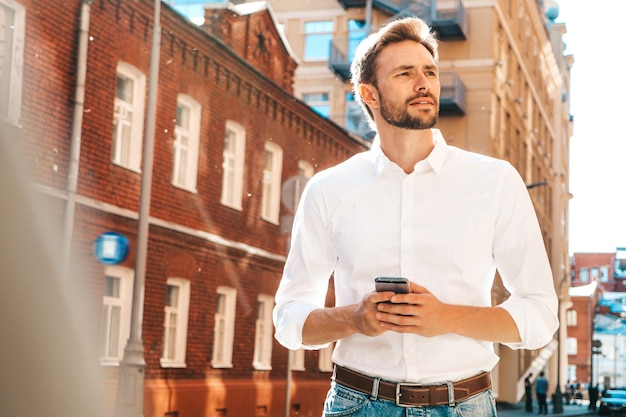 Bonito sorridente elegante hipster lambersexual modelo Homem moderno vestido com camisa branca Moda masculina posando no fundo da rua Ao ar livre ao pôr do sol Usando aplicativos de smartphone Segurando o celular