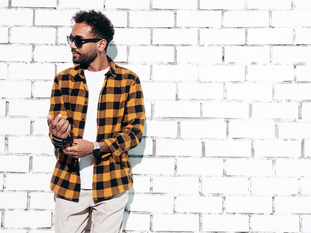 Foto bonito sorridente modelo hipster homem com barba sexy vestido com camisa amarela de verão e roupas jeans moda masculina posando perto da parede de tijolos brancos no estúdio em dia ensolarado isolado em óculos de sol