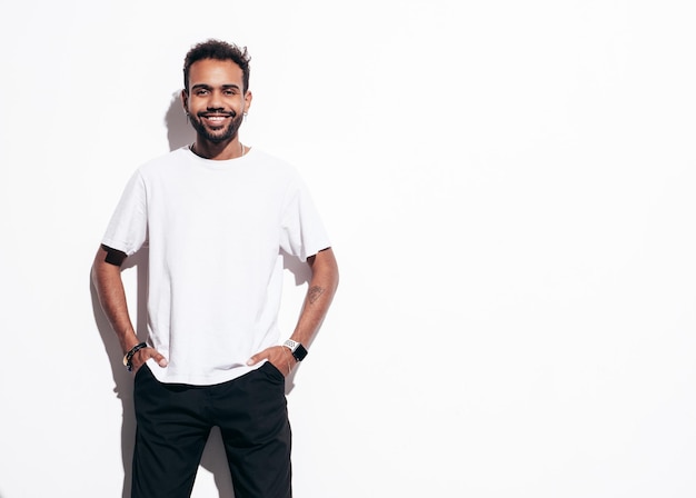 Bonito sorridente modelo hipster homem com barba sexy vestido com roupas jeans e camiseta de verão moda masculina posando perto da parede branca no estúdio em dia ensolarado isolado