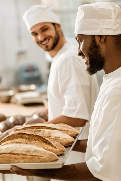 Bonitos jovens padeiros de chapéu na fabricação de panificação olhando pães frescos na bandeja