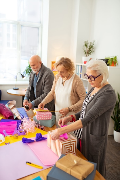 Bonitos regalos. Agradables personas mayores de pie en la habitación mientras preparan regalos para sus amigos