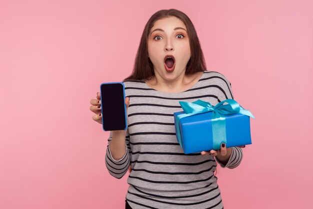 Bônus de presente de feriado inacreditável para usuário móvel Retrato de mulher chocada espantada segurando caixa de presente e telefone celular com display vazio para publicidade de compras on-line tiro de estúdio interno isolado