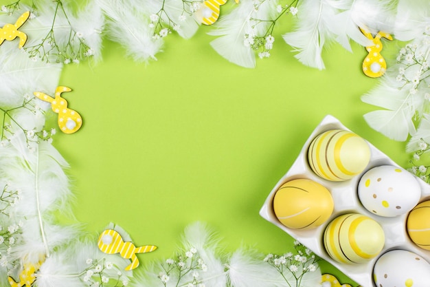 Borde de Pascua plano con huevos de colores en un soporte de cerámica blanca flores de primavera conejitos amarillos y plumas blancas sobre un fondo verde Vista superior Espacio de copia