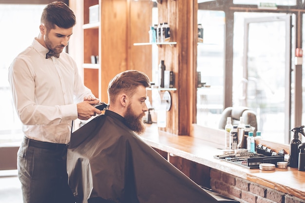 Bordes afilados. Vista lateral del joven barbudo cortándose el pelo por peluquero con maquinilla de afeitar eléctrica mientras está sentado en una silla en la barbería