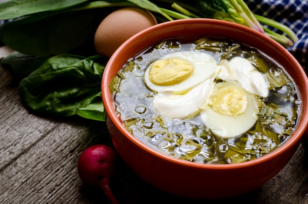 Borsch verde com azedas, espinafre e batata, coberto com ovo cozido e creme de leite em uma mesa de madeira vintage.