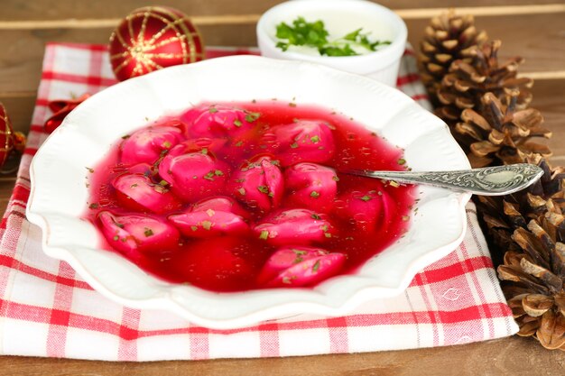 Borscht rojo claro polaco tradicional con bolas de masa en un recipiente en la bandeja y adornos navideños en la superficie de madera