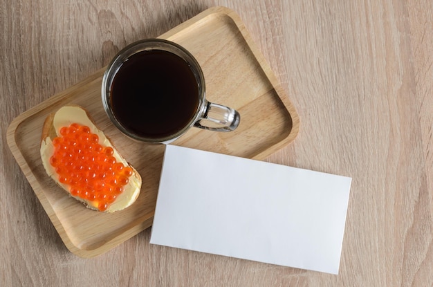 Foto bosquejo. un sándwich con mantequilla y caviar rojo con una taza de café negro caliente en una bandeja de madera. enfoque selectivo.