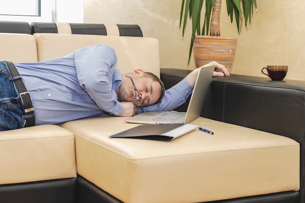 Boss Geschäftsmann in einem blauen Hemd ist auf der Couch vor einem Laptop eingeschlafen. Erschöpfter Mann, der sich im Büro entspannt. Die verantwortliche Führungskraft ist bei der Arbeit eingeschlafen.