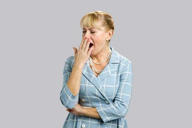 Bostezo mujer madura en gris. Retrato de mujer madura atractiva con expresión facial somnoliento, cansado, soñoliento, bostezando detrás de la mano.