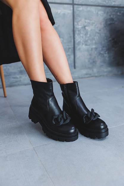 Botas de cuero negro sin tacones con hebilla piernas femeninas delgadas y una silla de madera sobre un fondo gris El concepto de una nueva colección de zapatos para la primavera Mujer con un vestido negro con una hendidura