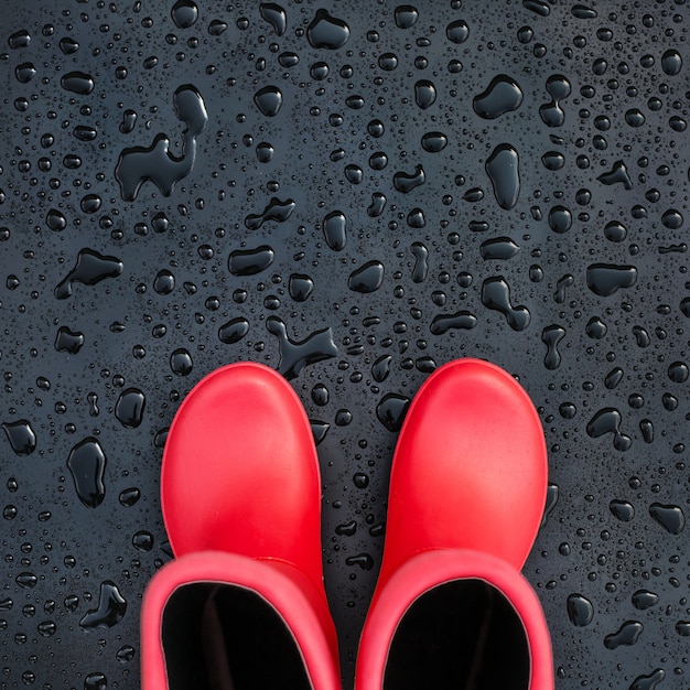 Botas de borracha vermelhas na moda na superfície molhada preta coberta com os pingos de chuva.