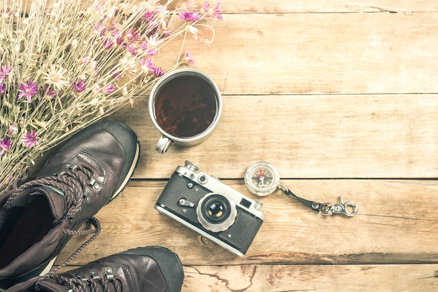 Botas, flores silvestres, una copa de metal, una brújula y otros atributos para una caminata en una superficie de madera. Concepto de senderismo en las montañas o el bosque, turismo, tienda de campaña, campamento. Vista plana, vista superior.