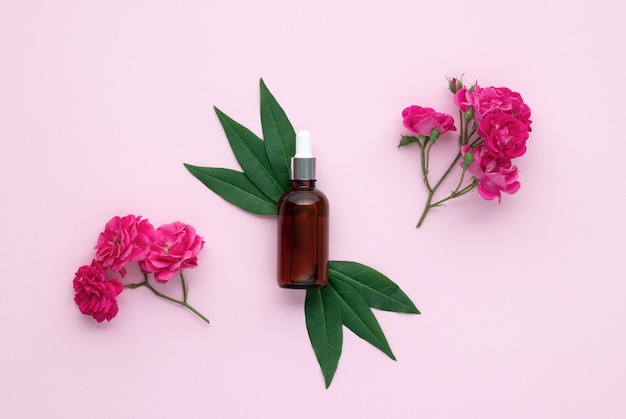 Botella de aceite esencial de rosa para masaje facial Vial de vidrio y hojas verdes en papel rosa fondo vista superior fragancia floral composición plana con flores producto de belleza natural para el cuidado de la piel