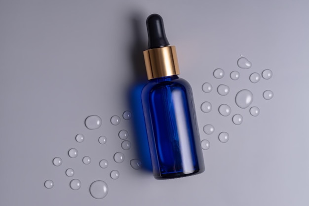 Botella de aceite esencial de vidrio azul en blanco con pipeta sobre fondo gris decorada con gotas de agua Concepto de cuidado de la piel con cosméticos naturales