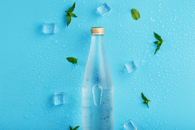 Botella de agua fría, cubitos de hielo, gotas y hojas de menta en azul.