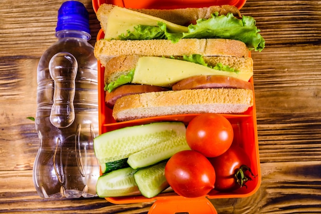 Botella de agua y lonchera con sándwiches, pepinos y tomates en la mesa de madera Vista superior