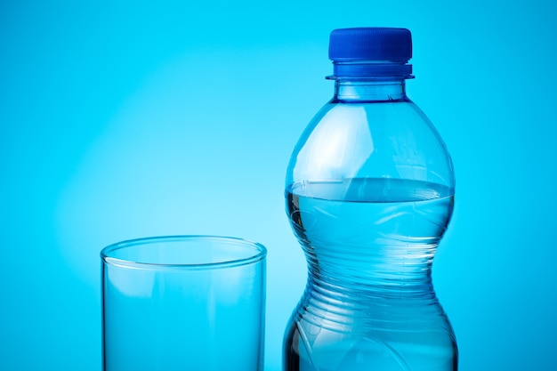 Una botella de agua mineral y un vaso vacío sobre un fondo azul.