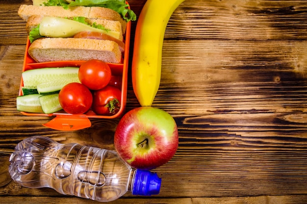 Botella de agua de plátano de manzana madura y lonchera con sándwiches de pepinos y tomates en la mesa de madera Vista superior