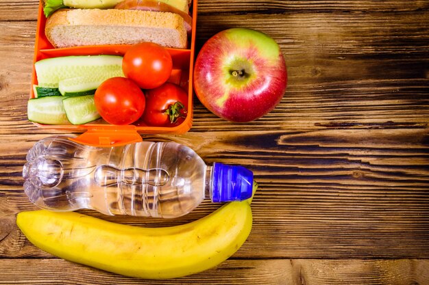 Botella de agua de plátano de manzana madura y lonchera con sándwiches de pepinos y tomates en la mesa de madera Vista superior