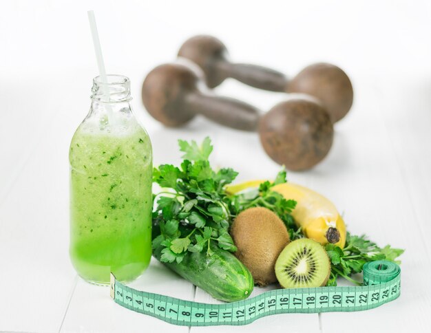 Una botella de batidos de pepino, fruta y cinta métrica sobre una mesa blanca.