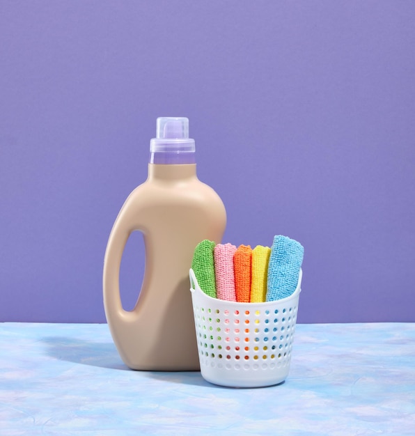 Una botella beige de detergente de lavandería líquido y una canasta blanca con un conjunto de toallas pequeñas de colores en un fondo púrpura