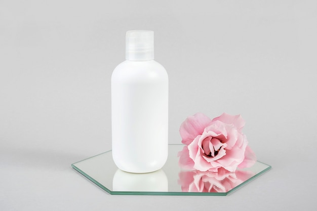 Botella en blanco cosmética blanca y flor rosa en el espejo, fondo gris. Maqueta de concepto de belleza cosmética de spa orgánico natural, vista frontal.