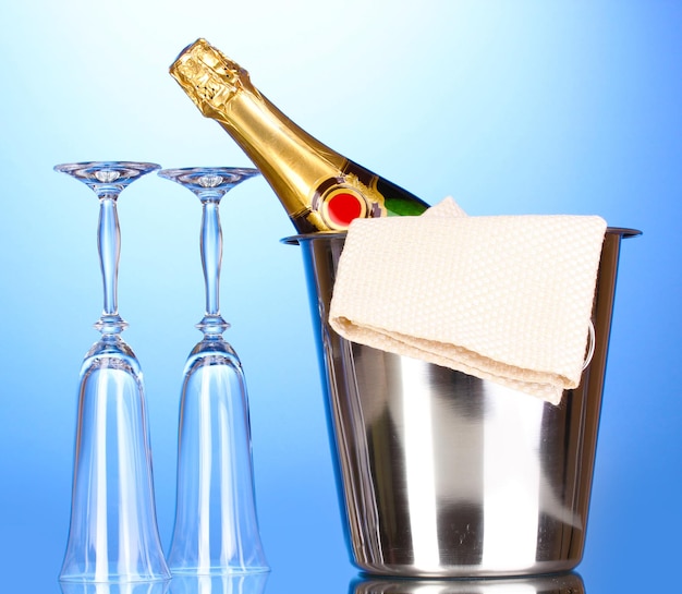 Botella de champán en un balde con hielo y vasos sobre fondo azul.