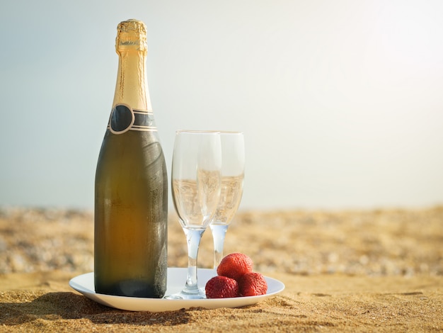 Una botella de copas de champán frío y fresas en una playa de arena.