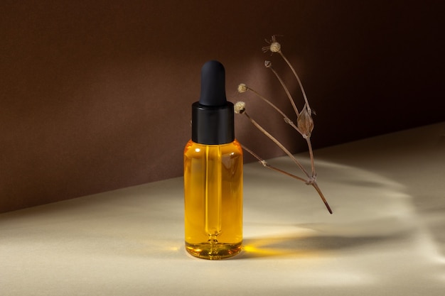 Una botella cosmética de vidrio con un gotero se encuentra junto a una flor seca sobre un fondo beige a la luz del sol. El concepto de cosmética natural, aceite esencial natural.