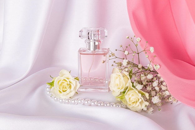 Una botella elegante de perfume o agua de baño para mujeres sobre un fondo de satén blanco y una bufanda de chiffon rosa flores blancas cuentas de perlas naturaleza muerta