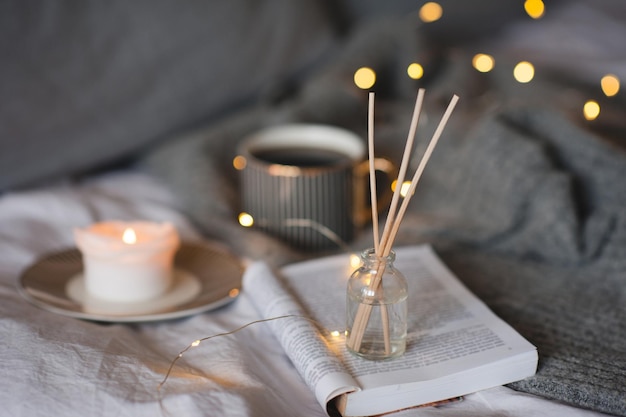 Botella con fragancia casera líquida y palos de bambú de madera, velas que queman scentes y una taza de té negro sobre luces incandescentes en el fondo de cerca. Temporada de vacaciones de invierno. Ambiente hogareño romántico y acogedor.