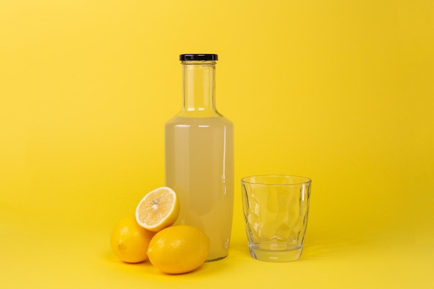 Botella con jugo de limón y un vaso vacío al lado y limones enteros y en rodajas aislados en un fondo amarillo