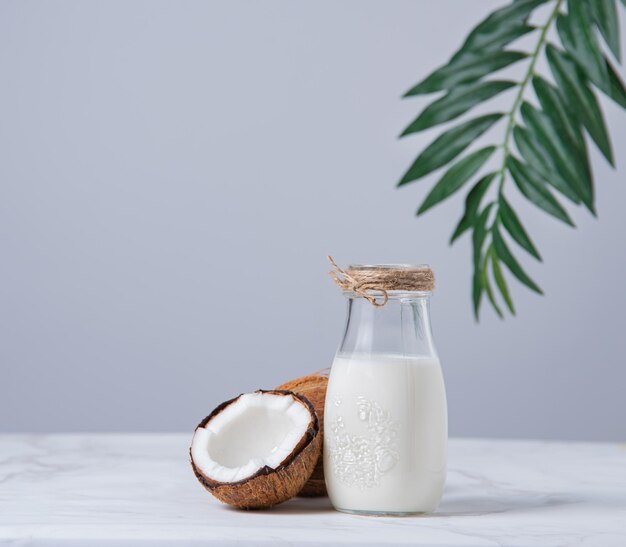 Botella de leche vegana de vidrio sobre mesa de mármol blanco con medio cocos y rama de palma. Vista frontal y copia de la imagen del espacio.
